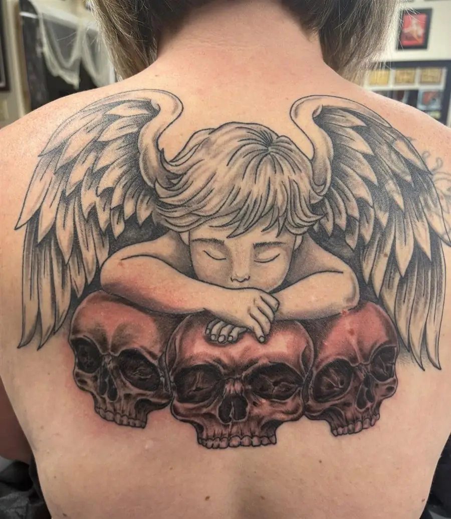 Black and grey cherub angel resting on skulls upper back tattoo