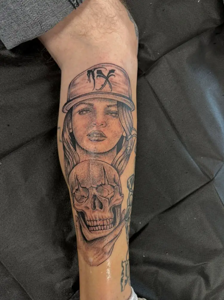 Black and grey portrait of a girl in a TX cap holding a skull, forearm tattoo